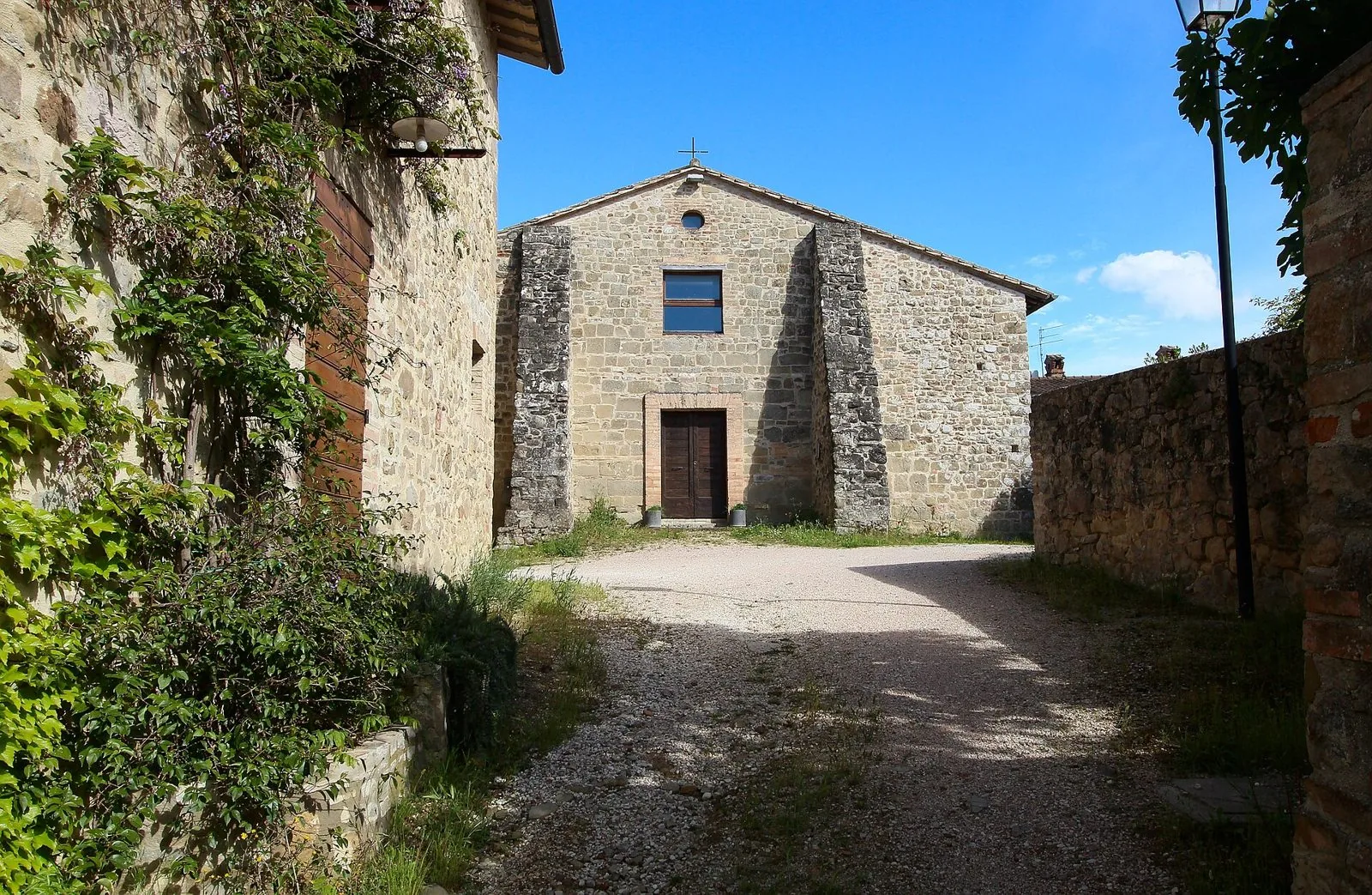 Facciata in pietra della chiesa del complesso di San Lorenzo a Collazzone, con viottolo di ghiaia e muri coperti di rampicanti