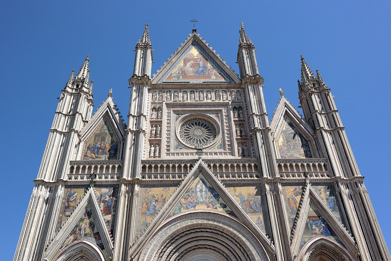 La facciata gotica del Duomo di Orvieto vista dal basso, con guglie, mosaici dorati, statue e il rosone centrale contro il cielo azzurro